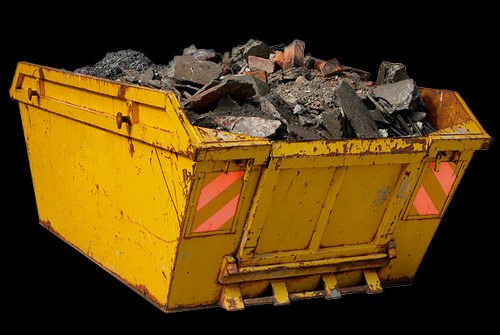 A large, yellow industrial skip positioned on a flat surface, filled with irregularly shaped debris including broken bricks, concrete chunks, and plaster, predominantly in shades of grey, brown, and reddish hues. The exterior of the skip shows visible signs of wear with scratches and stains, and it features reflective pink and orange safety markings on its rear panels. The skip's metal surface has a textured finish and is equipped with a reinforcement bar along the lower edge for durability. The background is completely black, isolating the skip and emphasizing the payload of construction or renovation waste, consistent with rubbish removal services offered by Commercial Waste Heston in the Heston area or nearby postcode regions.