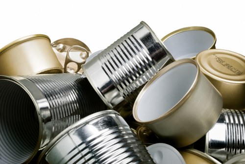 A close-up image depicting a collection of metal food cans piled together, including both opened and unopened tins, with some having pull-tab lids partly visible. The cans vary in size and are primarily silver, with a few having gold or metallic finishes, displaying a mixture of smooth and ridged textures. They are arranged randomly, with some lying on their sides and others upright, set against a plain, light-colored background. The image highlights the reflective surfaces of the cans, emphasizing their metallic nature, and suggests an environment related to waste or recycling collection, which aligns with rubbish removal services in the Heston area. The clear focus on the cans indicates the removal or disposal of metal packaging as part of waste management tasks performed by Commercial Waste Heston, capturing the typical scene of cardboard and metal waste associated with commercial or household rubbish collection in the local area.