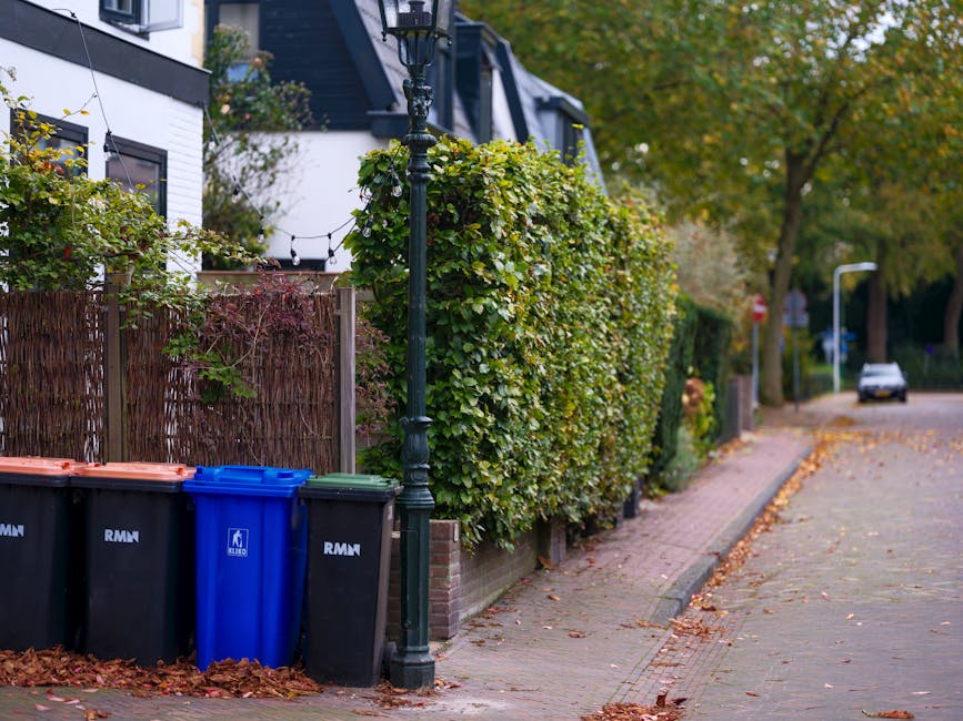 An outdoor residential scene showing four wheelie bins positioned on a paved sidewalk next to a wooden fence and a tall, black decorative street lamp. The bins include two black containers with orange