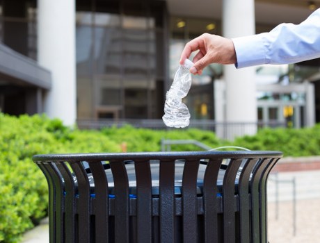A person wearing a light blue long-sleeved shirt is seen disposing of a crumpled clear plastic water bottle into a black plastic outdoor rubbish bin with vertical slats, situated on a paved area near a modern commercial building with large windows and metallic framing. The container is positioned on a paved surface with a low green hedge in the background, indicating a tidy external environment consistent with commercial waste management practices in Heston. The image emphasizes proper waste disposal of plastic materials, supporting rubbish removal services offered by Commercial Waste Heston in the local area, with natural daylight illuminating the scene and highlighting the textured surface of the plastic bottle and the open top of the bin.
