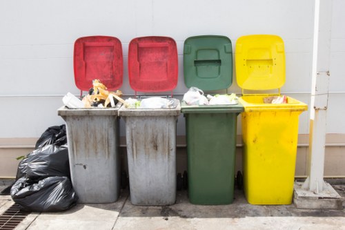 A row of four wheelie bins positioned outdoors on a paved surface against a white wall, with their lids open and filled with various waste items. From left to right, the first grey bin contains black plastic bags and some loose waste, its lid partially covering the contents. The second grey bin also holds waste bags and miscellaneous debris visible from its open lid. The third green bin has a closed lid, but some waste is spilling slightly over the edge, with paper or packaging materials visible. The fourth yellow bin, also with its lid open, appears to contain paper, plastic, and other rubbish. To the left of the bins, there are additional black plastic bags piled on the ground, likely for refuse collection or disposal. The scene suggests a typical refuse disposal area, commonly serviced by waste management providers like Commercial Waste Heston for rubbish removal services in the Heston area, within postcode TW5, offering solutions for commercial waste collection and rubbish disposal.