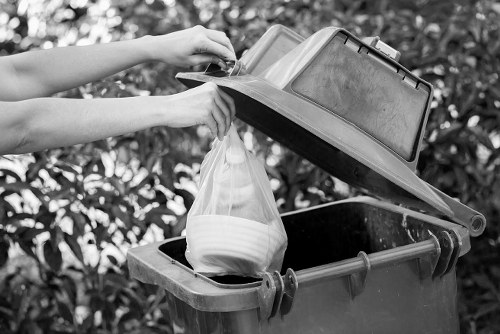 A person is seen disposing of waste by placing a plastic bag filled with white crockery into a large, dark grey rubbish bin situated outdoors on a paved driveway. The bin has a partially open lid, revealing a spacious interior, and is positioned against a background of leafy shrubs, indicating a residential or commercial property grounds. The person's arms are visible, showing them lifting the lid with one hand while inserting the bag with the other. The plastic bag appears semi-transparent, and the crockery inside is white, contrasting with the dark interior of the bin. The scene is lit by natural daylight, capturing the textures of the plastic bag, the smooth surface of the crockery, and the rough finish of the bin. This image relates to rubbish collection and waste disposal services, exemplified by Commercial Waste Heston, serving the Heston area close to postcode TW5, highlighting practical waste management activities typical of the local environment.