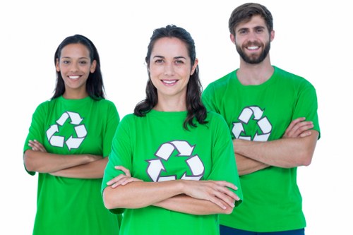 A close-up group photograph of three individuals standing side by side outdoors, wearing bright green T-shirts featuring a large white recycling symbol. The woman in the center, with shoulder-length dark hair, is smiling directly at the camera with her arms crossed. To her left, a woman with long black hair is also smiling and has her arms crossed, while to her right, a man with brown hair and a beard is smiling with his arms crossed as well. The background is blurred and suggests an outdoor environment, with natural lighting highlighting their cheerful expressions and casual pose, emphasizing a positive attitude towards waste management and recycling efforts in the context of rubbish removal services, such as those offered by Commercial Waste Heston.